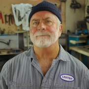 Man with gray hair and beard, wearing coveralls and standing in a workshop